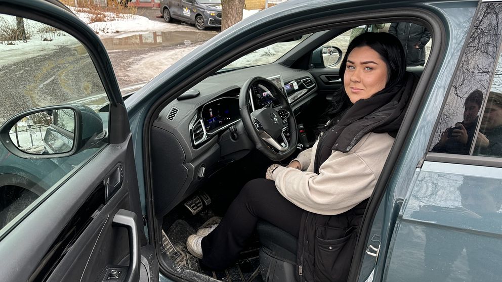 Person sitter i förarsätet på en bil med öppen dörr utomhus.