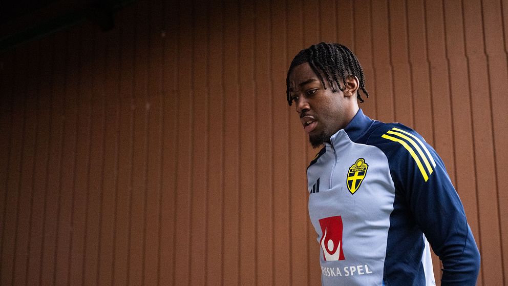 En fotbollsspelare i träningskläder promenerar vid trävägg.