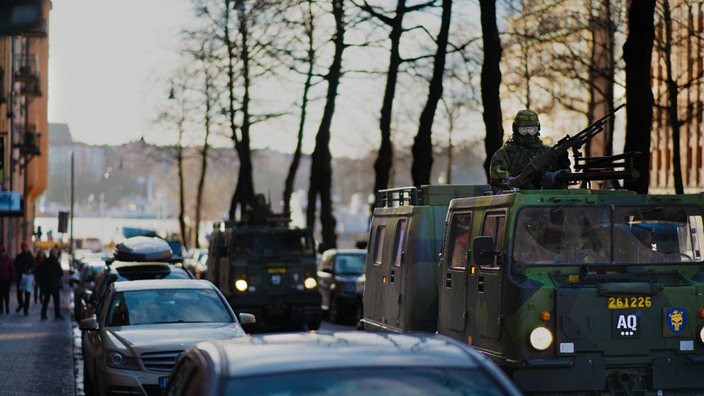 Militära fordon kör genom en stadsgata kantad av parkerade bilar som omges av träd. En soldat sitter på taket av ett av fordonen med en kulspruta, och flera soldater verkar finnas i de militära fordonen i kolonnen. Scenen äger rum under en sen eftermiddag med ett mjukt ljus i bakgrunden.