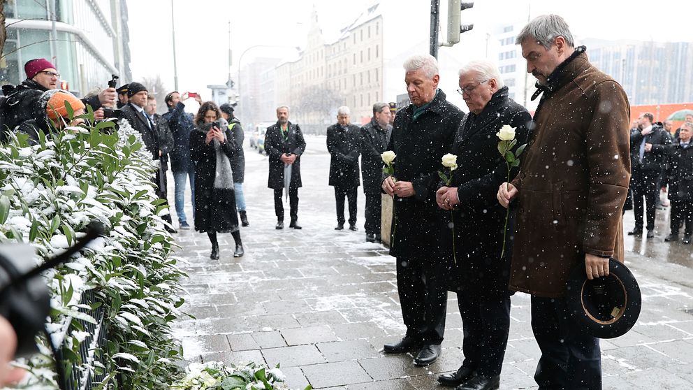 Människor deltar i en minnesceremoni vid en plats täckt med blommor under en snöig dag. Tre män håller vita rosor framför dem medan andra deltagare och åskådare står i bakgrunden.