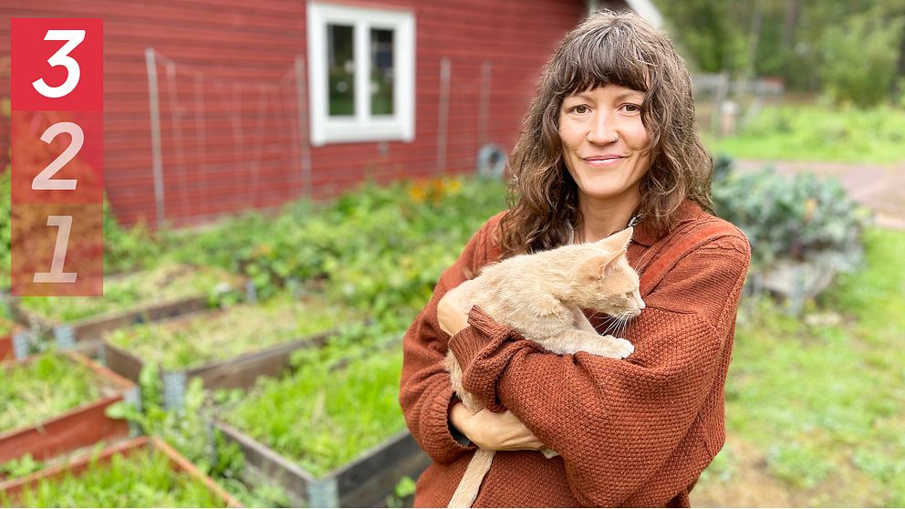 kvinna håller i katt, står framför odlingar vid rött trähus