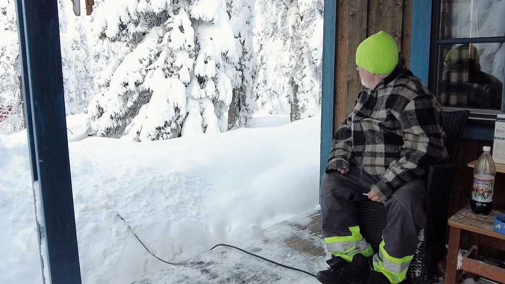 En man i neongrön mössa sitter på en altan till en stuga i Sälen och tittar ut på ett tjockt snötäcke.