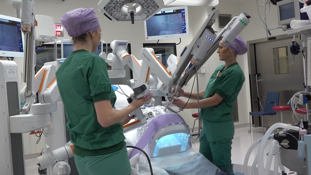 Två sjukvårdspersonal klädda i gröna operationskläder står vid en avancerad kirurgisk robot i ett operationsrum. Den ena personen håller i en kontroll och fokuserar på robotens instrument, medan den andra justerar maskinens position. I bakgrunden syns flera skärmar som visar olika medicinska data och bilder. Rummet är modernt och välutrustat för kirurgiska ingrepp.