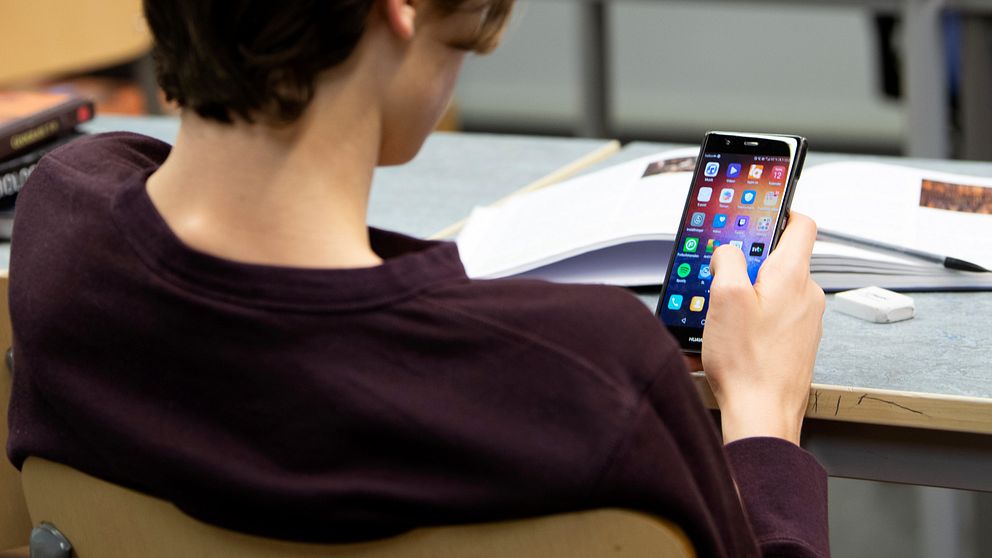 Ung kille sitter med mobiltelefon i en skolsal.