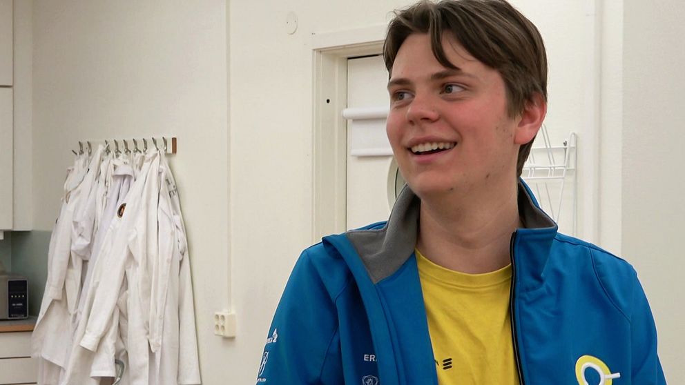 Person i blå jacka och gul tröja står i ett rum med laboratorierockar på väggen.