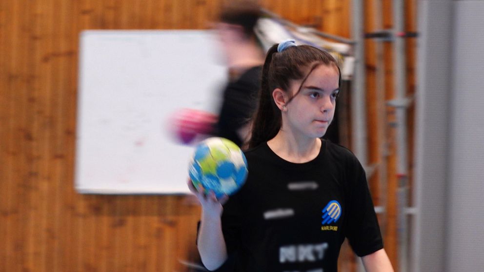Handbollsspelare med boll i handen under en inomhusträning. Trävägg och whiteboard i bakgrunden.