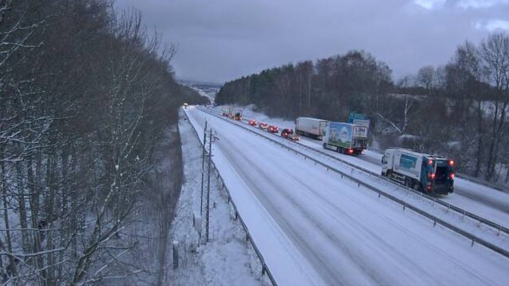 Ett snöigt landskap längs en motorväg med flera fordon, inklusive lastbilar och en sopbil. Träden vid vägen är täckta av snö, och himlen är grå och molnig, vilket skapar en vintrig och kall atmosfär.