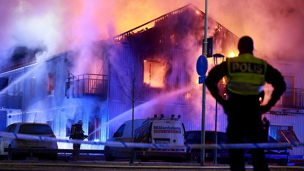 En polis står vid avspärrningarna och ser på när brandmän bekämpar en stor brand i en byggnad i Hölö under natten. Flammor och rök väller ut från det eldsvåda huset, med blå och röda ljus som reflekteras från räddningstjänstens fordon.