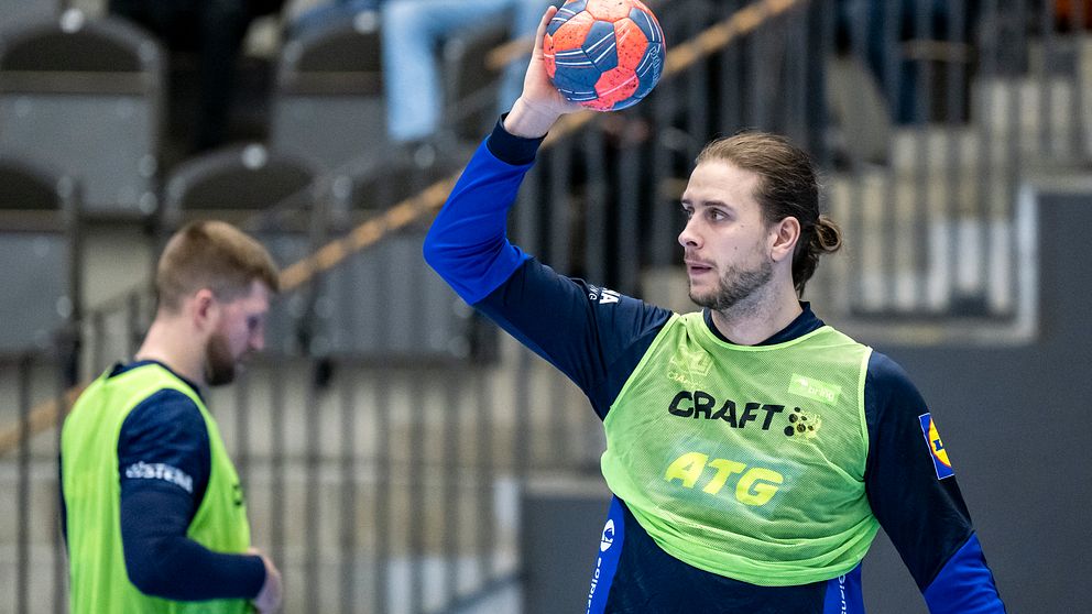 En handbollsspelare i en grön träningströja förbereder sig för att kasta en handboll, medan en annan spelare står i bakgrunden. Arenan har åskådare i bakgrunden och ger en känsla av en aktiv sportmiljö.