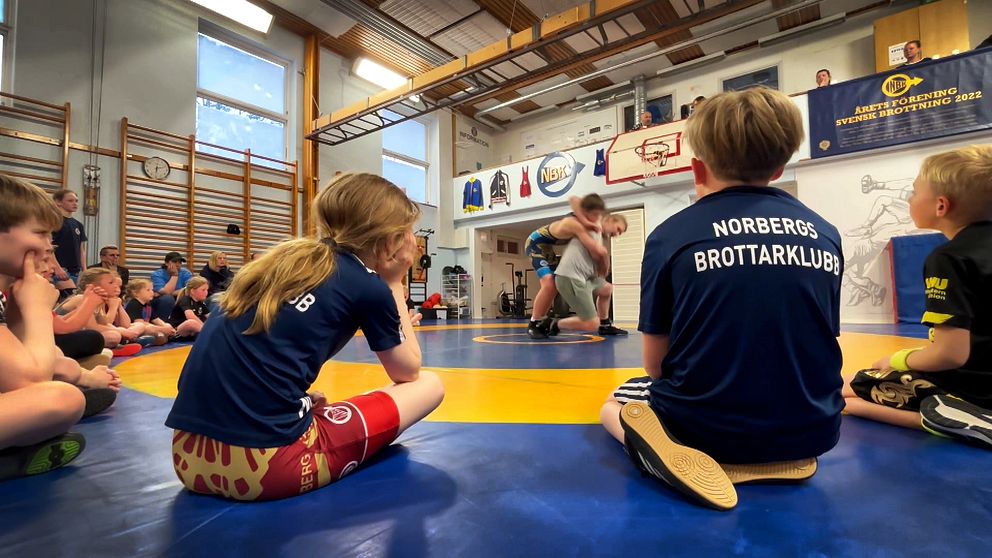Barn i idrottshall sitter på mattan och tittar på en brottningsmatch.