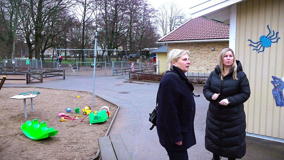 Två personer står på en gård vid en förskola, med en lekplats i bakgrunden som innehåller gungor och sandlådor. Det finns leksaker som ligger på marken, inklusive en grön rutschkana och andra färgglada leksaker. Byggnaden har en väggmålad dekoration av en spindel och det är molnigt ute.