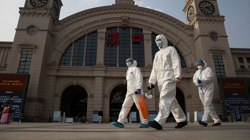 Människor klädda i skyddsdräkter och ansiktsmasker går framför en historisk byggnad med klocktorn, i Wuhan, Kina, under en presumtiv saneringsinsats.