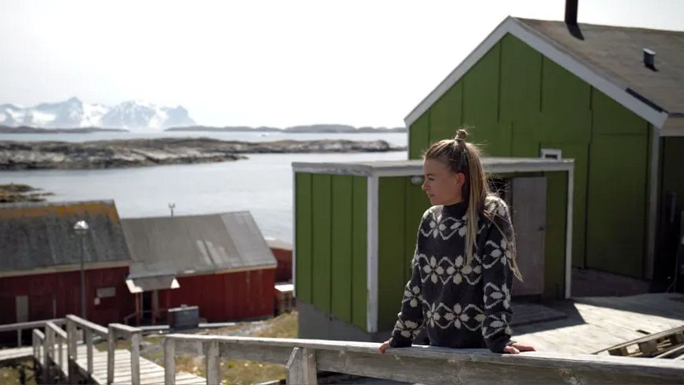 Kvinna i mönstrad tröja blickar ut över traditionella hus vid kusten i en nordlig miljö.