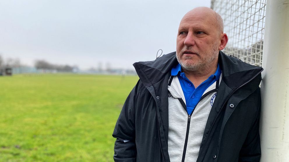 Peter Upphoff, distriktschef för Hallands fotbollförbund står lutad mot ett stängsel vid Alevallen i Halmstad. Han bär en svart jacka över en grå tröja och har en blå skjorta under. I bakgrunden syns en stor grön yta och en suddig himmel.