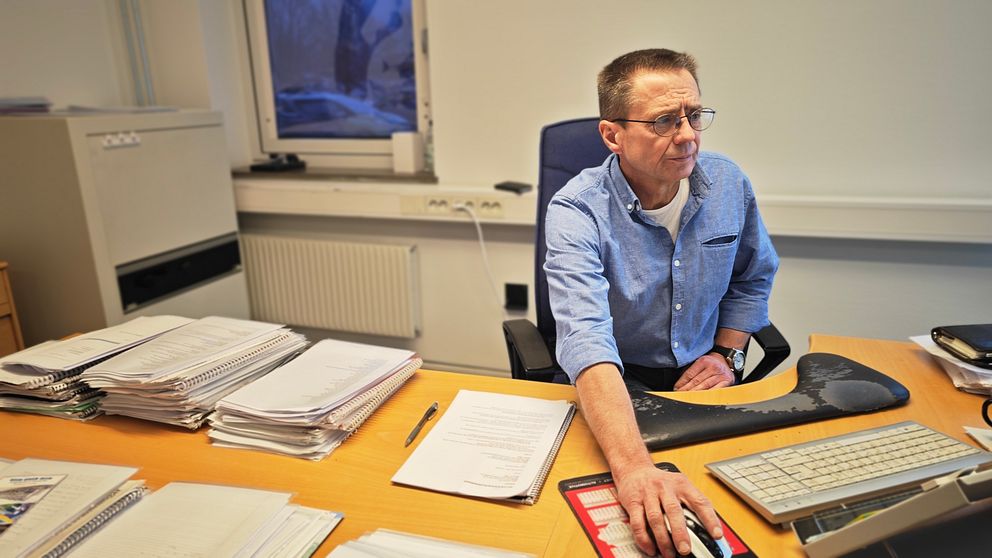 En man sitter vid ett skrivbord med många pappershögar. Han håller handen på en datormus och tittar mot en datorskärm