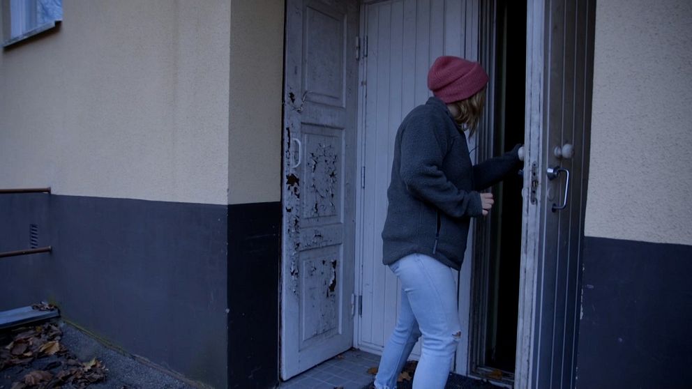 En person i en grå jacka och röd mössa står vid en dörr och förbereder sig att gå in i en byggnad. Dörren är delvis öppen och det finns en sliten vit dörr bredvid. Området har höstlöv på marken och byggnaden har en enkel, modern utformning.