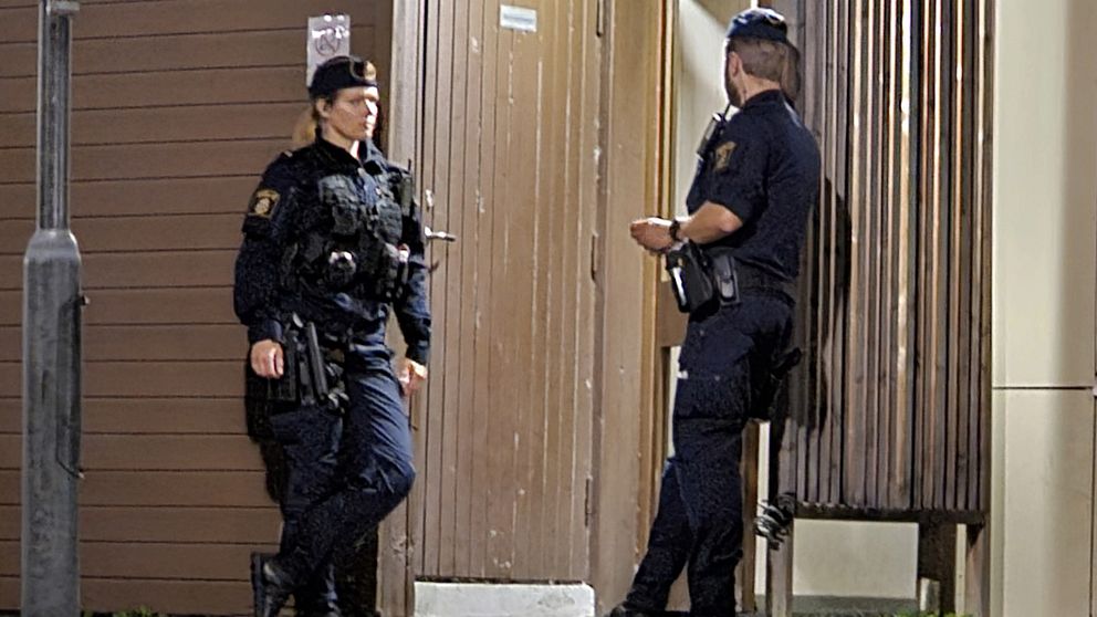 Två poliser i uniform står vid en dörr i en mörklagd miljö. Den ena polisman tittar på dörren medan den andra står lutad mot en vägg, båda med vapen synliga. Det är kväll och belysningen skapar en dramatisk stämning.