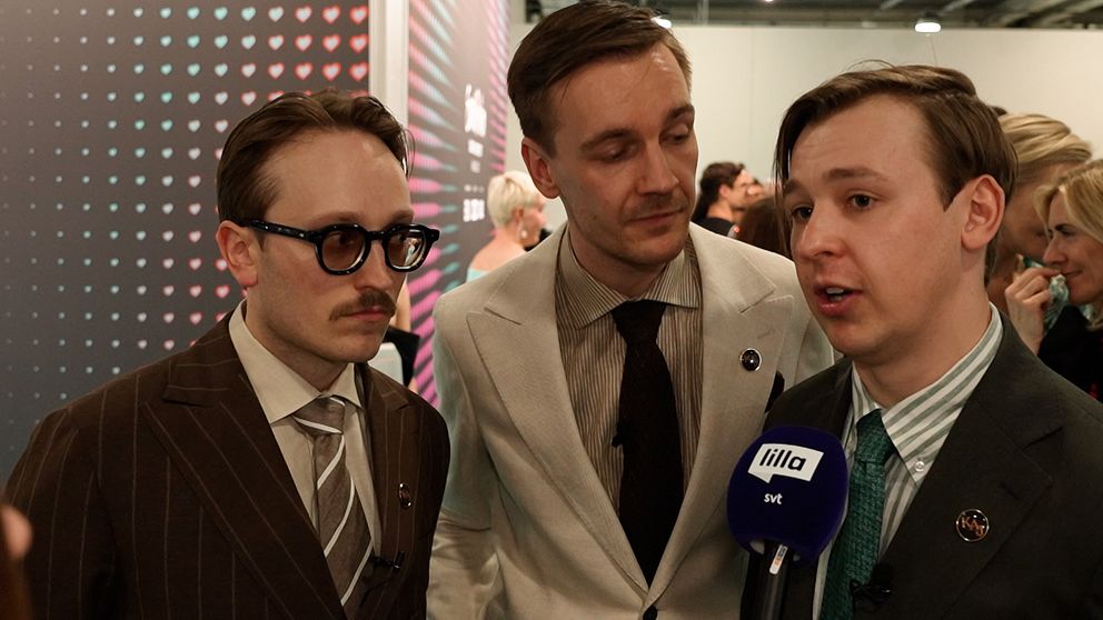 Tre personer i kostym intervjuas av media på ett evenemang, mikrofon med SVT-logga syns.