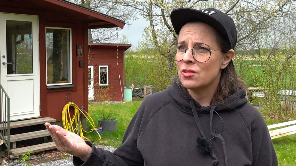 Person med keps utanför ett rött hus med trädgårdsslang och grönska i bakgrunden.