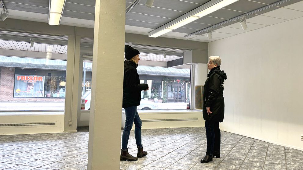 Två personer står och samtalar i en tom lokal med stora fönster som vetter mot en gata där en skylt med texten ”Frisör” syns.