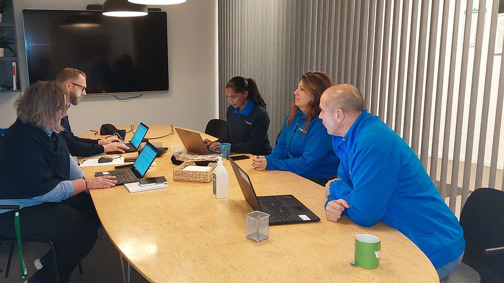 En grupp på fem personer deltar i ett möte i ett modernt konferensrum. De sitter vid ett stort träbord med laptops och anteckningar framför sig. I bakgrunden syns en stor bildskärm och en vägg med stående paneler. Rummet har en ljus och luftig känsla med växtlighet synlig i hyllorna. Alla deltagare för fokus på sina skärmar och verkar engagerade i diskussionen.