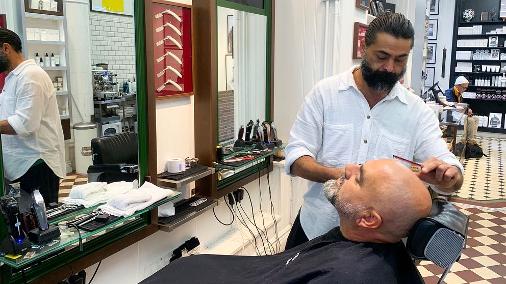 En man får en frisyrbehandling i en barberarsalong medan en annan barberare arbetar i bakgrunden. Salongen har en modern inredning med speglar och olika verktyg på en arbetsbänk. Golvet är mönstrat med rutor.