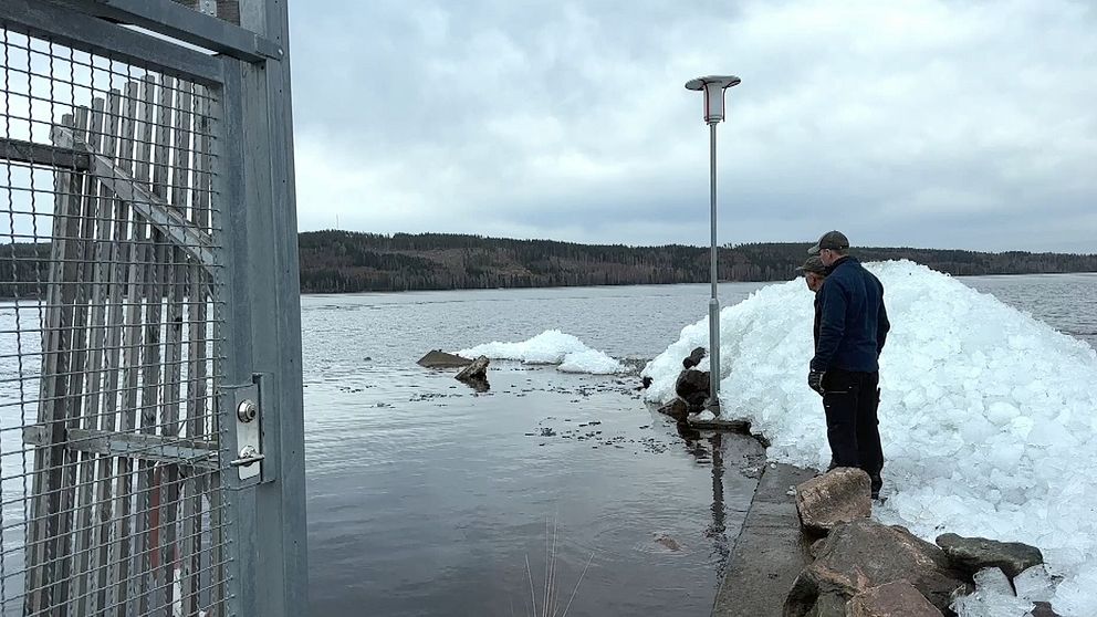 Två män står på en betongpir och blickar ut över sjön. Till höger har de en stor hög is, som ligger över betongpiren
