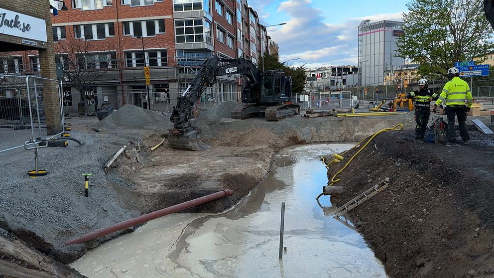 Grävmaskin och byggarbetare vid avspärrad grop på en stadsgata under arbete.