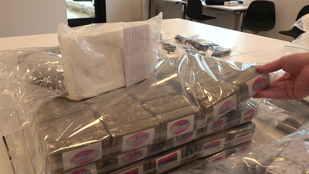 Stapel av beslagtagna droger inslagna i plast på ett bord, med en hand som håller i ett av paketen. I bakgrunden syns kontorsstolar och bord.