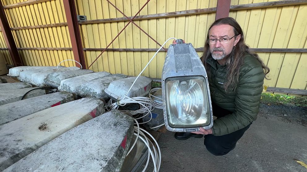 SVT:s reporter håller upp en gammal belysningsanordning framför en stapel av betongblock, som ser ut att ha kablar anslutna. Bakgrunden visar en trävägg i gult och rödmetalliska stänger som stöder strukturen.