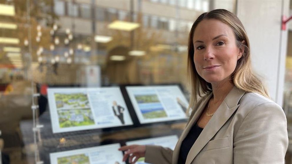 Mäklare Marie Ekängen står framför Länsförsäkringars skyltfönster och pekar på bilder av hus till salu. Tavlorna är organiserade i två sektioner, med både text och illustrationer som verkar relatera till landskap eller projekt. Bakgrunden visar en känsla av en affärsmiljö.