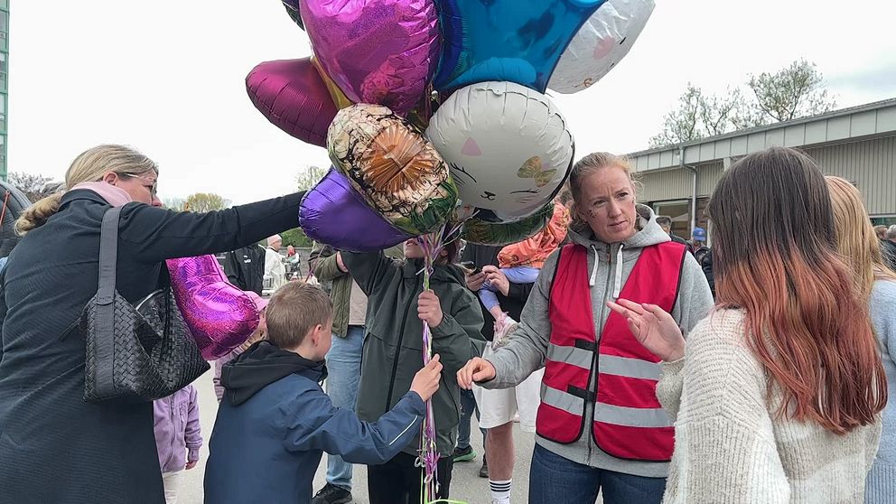 Person i röd väst delar ut heliumballonger till barn och vuxna vid en utomhusaktivitet.