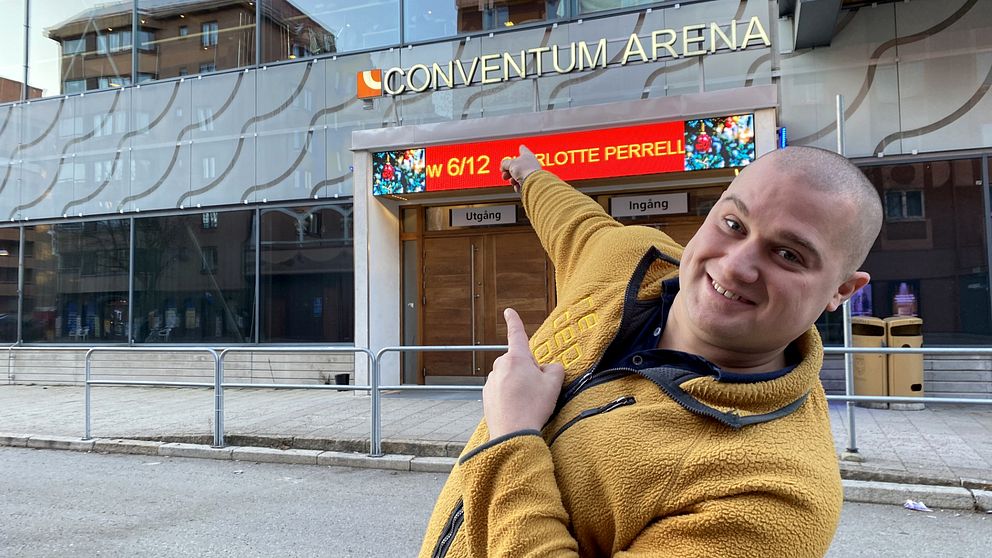 En man som ler och pekar på den stora arenabyggnaden Conventum i Örebro bakom sig.
