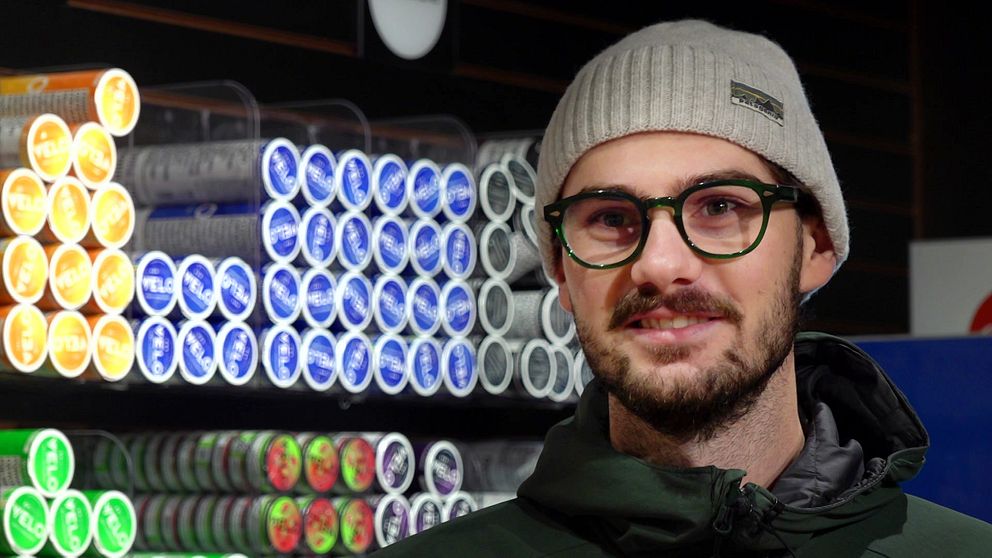En person står i en butik framför en hylla fylld med snusdosor av olika färger, inklusive gult, blått, grönt och rött.