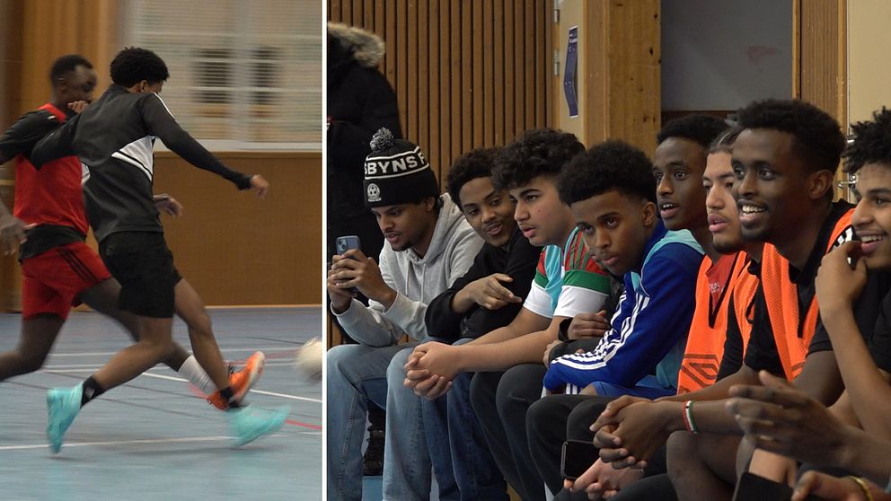 Två personer spelar fotboll inomhus, klädda i träningskläder.  En grupp personer sitter på en bänk,