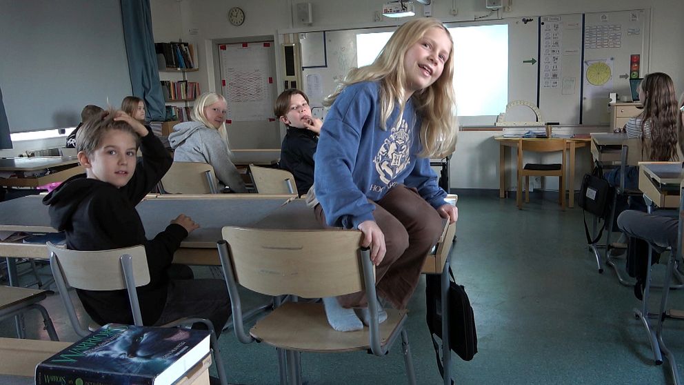 En klassrumsscen där en elev sitter på en stol och verkar delta i lektionen, medan andra elever sitter i sina bänkar. I bakgrunden syns en projektor och en grön tavla. Det finns skrivböcker och andra skolmaterial synliga på bänkarna.