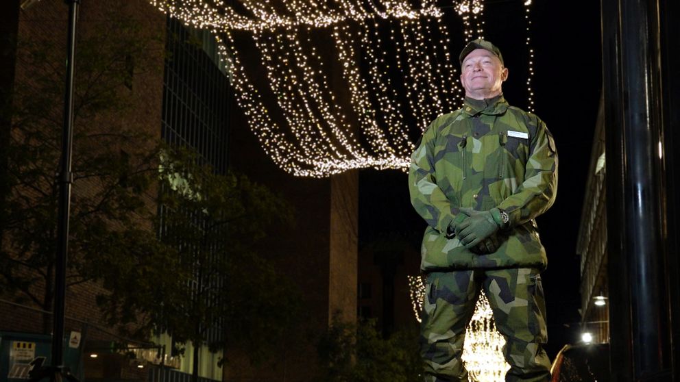 Lars Bergman i camouflagedräkt står uppställd på en gatan där julbelysning hänger ovanför. Omgivningen är mörk med ljusa dekorationer som skapar en festlig stämning.
