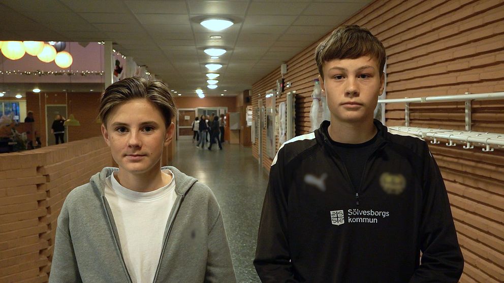 Två personer står i en lång korridor med träväggar och belysning i taket. Den ena personen bär en grå hoodie, medan den andra har på sig en svart tröja med logotyp. I bakgrunden syns flera människor som går längs korridoren. Mobilförbud i skolan