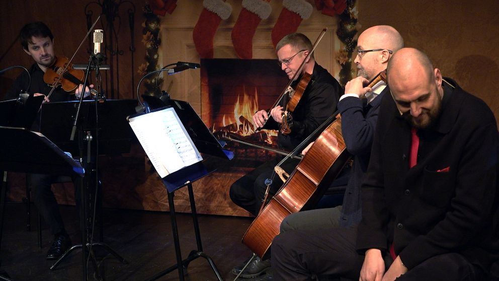 En musikensemble framför ett stycke i en mysig miljö med en julinspirerad bakgrund, inklusive en eldstad och julstrumpor. Instrument som violin och cello är synliga, och notställ med noter står framför musikerna. Atmosfären är varm och festlig.