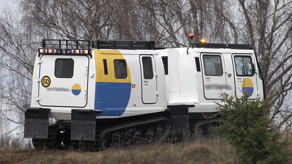 Fordonet är ett specialiserat terrängkörande fordon med banddrift, designat för tuffa väderförhållanden. Det har en vit kaross med gula och blå detaljer samt en tydlig nummerskylt, vilket tyder på att det används för specifika uppdrag, eventuellt inom transport eller inspektion. Bakom fordonet syns träd och en dyster himmel som bidrar till den allmänna atmosfären.