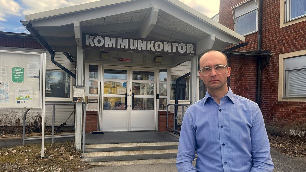 Bjurholms kommunchef Jimmy Johansson står i blå skjorta framför kommunkontoret med bister uppsyn.