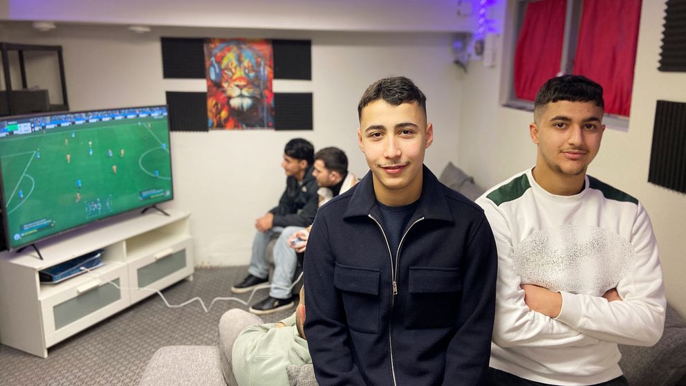 Abude Habbak och Hussein Hassani står framträdande i förgrunden, medan tre andra sitter bakom dem och spelar FC25 som visas på en TV. Rummet har en avslappnad atmosfär med en ljus dekor och mörka element på väggarna. En färgglad tavla hänger ovanför TV:n.