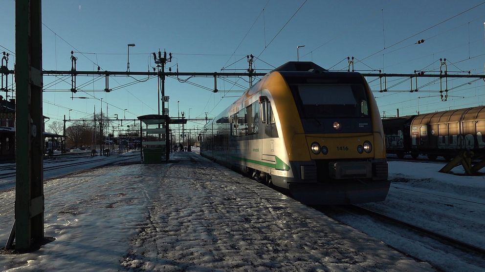 Tåget ankommer till en tågstation täckt av snö. Tåget är gult och grönt, och stationens plattform syns i förgrunden, med en busshållplats till vänster. I bakgrunden finns flera järnvägsspår och en klar blå himmel.