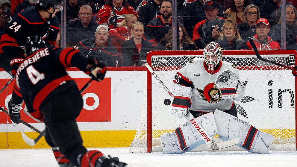 Spelare från två hockeylag är i aktion på isen. En spelare i röd tröja och nummer 24 skottar pucken mot målet, medan målvakten i vit och svart utrustning, nummer 35, förbereder sig för att med hjälp av utrustningen stoppa pucken. I bakgrunden syns åskådare som hejar på.