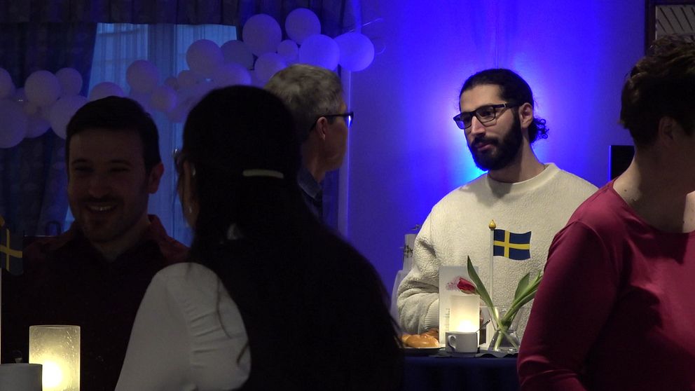 Hela scenen visar en social sammankomst med personer som interagerar i en festlig miljö. Flera personer står och pratar vid ett bord med en blomma och en svensk flagga på.