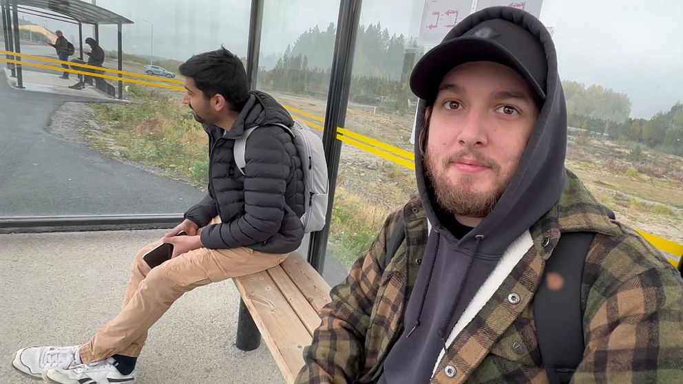 Två personer sitter vid en busshållplats inomhus. Den ena personen sitter på en bänk, klädd i en svart jacka och har en ryggsäck, medan den andra personen står bredvid, i en rutig jacka och hoodie. Området verkar vara dimmigt med grönska i bakgrunden.