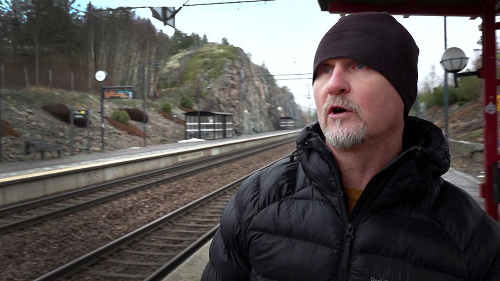 En person i en svart jacka står på en tågstation, med en tågperrong och berg i bakgrunden.