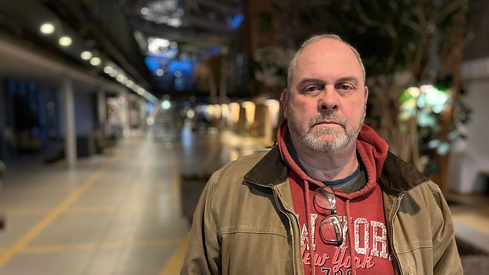 En person står i en inomhusmiljö, klädd i en khaki-jacka och en röd huvtröja med tryck. I bakgrunden syns en gångväg med belysning och växter