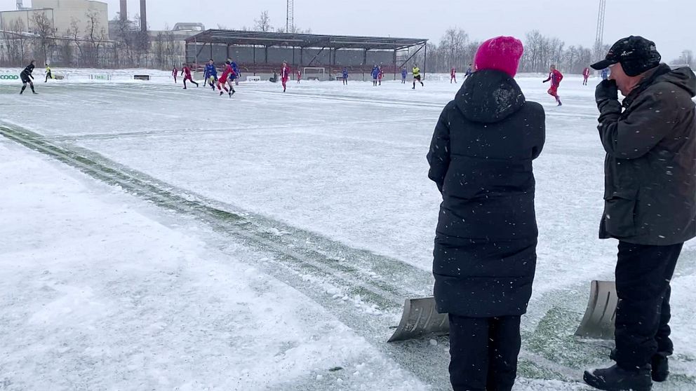 Personer ser på en fotbollsmatch under vintriga förhållanden på en snötäckt plan.