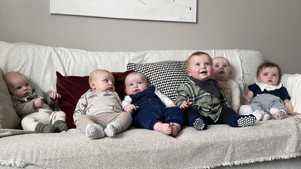 Sex bebisar sitter på rad i en vit soffa.